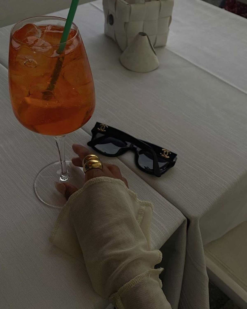 hand wearing gold rings holding a cocktail next to black sunglasses 
